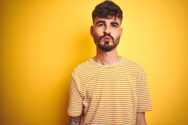 Çizgili tişörtü olan genç bir adam izole edilmiş sarı arka planda dikilip kameraya bakarak güzel ve seksi bir öpücük yolluyor. Aşk ifadesi.