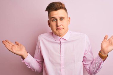 Soyutlanmış pembe arka plan üzerinde duran zarif gömlekli genç yakışıklı işadamı elleri ve kolları havada, kafası karışmış bir ifade takınıyor. Şüphe kavramı.