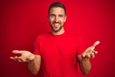 Kırmızı izole edilmiş arka plan üzerine gündelik tişört giyen genç yakışıklı adam gülümseyerek yardım eli uzatıyor ve kabul ediyor..