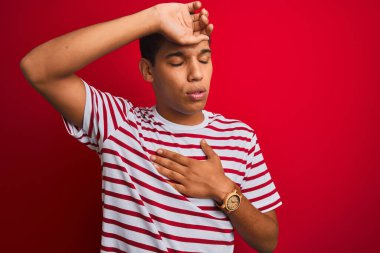 İzole edilmiş kırmızı arka plan üzerinde çizgili tişört giyen genç yakışıklı Arap adam hastalık, ateş ve soğuk algınlığı için alnına dokunuyor.