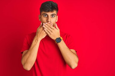 İzole edilmiş kırmızı arka plan üzerinde duran genç Hintli adam yanlışlıkla elleri ile ağzını kapatırken şok oldu. Gizli konsept.