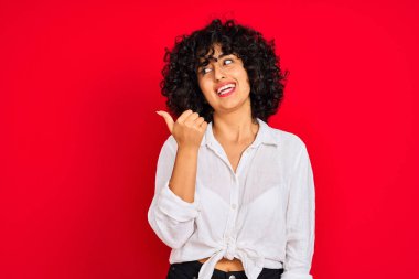 Kıvırcık saçlı, beyaz gömlekli, izole edilmiş kırmızı arka planda gülümseyen, gülümseyen ve baş parmağıyla yüzünü gösteren genç bir Arap kadın..