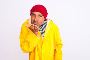 Yağmurluk giyen ve yünlü şapkalı orta yaşlı bir adam izole edilmiş beyaz arka planda dikilip kameraya bakarak havada öpücükler savuruyor. Güzel ve seksi. Aşk ifadesi.