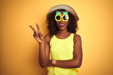 İzole edilmiş sarı arka planda komik ananas güneş gözlüğü takan Afro-Amerikan kadın kameraya gülümseyen ve zafer işareti yapan. İki numara..