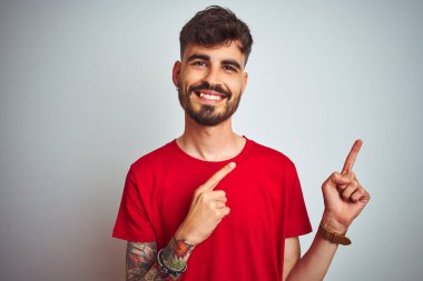 Kırmızı tişörtlü genç adam izole edilmiş beyaz arka plan üzerinde duruyor gülümsüyor ve kameraya bakıyor iki eliyle ve parmaklarıyla kenarı işaret ediyor..