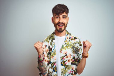 Tatilde dövmeli bir adam, izole edilmiş beyaz arka plan üzerinde durmuş kollarını kaldırarak ve açık gözlerle başarıyı kutluyor ve şaşırıyor. Kazanan konsept.