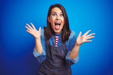 Genç ve güzel bir iş kadını mavi izole arka planda önlük giyip kollarını kaldırıp heyecanla haykırarak çılgınlığı ve başarıyı kutluyor. Kazanan konsept