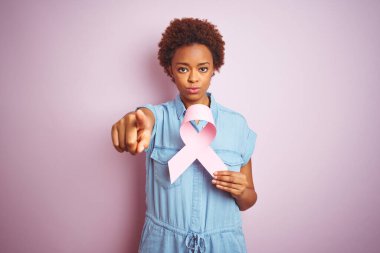 Genç Afro-Amerikalı kadın elinde Brest Kanser kurdelesi ile pembe arka planda parmağıyla kamerayı işaret ediyor ve sen, el işareti, ön taraftan pozitif ve kendinden emin bir jest.