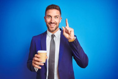 Genç yakışıklı iş adamı mavi bir arka plan üzerinde kahve içiyor. Bir fikir ya da soru işareti ile şaşırmış, mutlu yüzle bir numara.