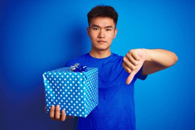 Asyalı genç bir Çinli 'nin elinde doğum günü hediyesi var. İzole edilmiş mavi arka planda duruyor. Kızgın bir yüzü var.