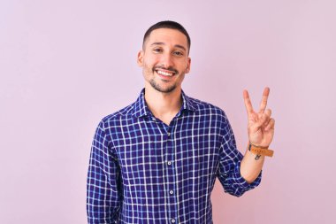 İzole edilmiş arka planda duran yakışıklı genç adam kameraya bakıp gülümsüyor ve zafer işareti yapıyor. İki numara..