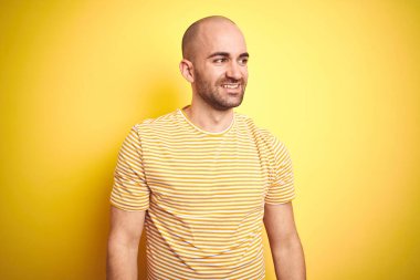 Sakallı genç ve kel bir adam sarı izole edilmiş arka plan üzerine günlük çizgili tişört giyiyordu. Yüzünde gülümseme ve doğal bir ifadeyle diğer tarafa bakıyordu. Kendinden emin gülen.