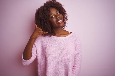 İzole edilmiş pembe arka planda duran kazak giyen genç Afrikalı kadın telefonla el ve parmaklarıyla telefonla konuşur gibi gülümsüyor. İletişim kavramları.