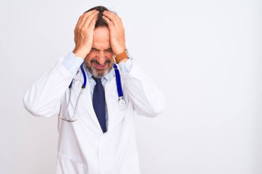 Paltolu ve steteskoplu orta yaşlı bir doktor, izole edilmiş beyaz arka planda dikiliyor. Ağrı ve migrenden dolayı baş ağrısı ve stresten muzdarip. Ellerini başının üstüne koy..