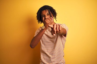 Rastalı tişörtlü Afro adam izole edilmiş sarı arka planda dikilip sana gülüyor, eli ağzında kamerayı işaret ediyor, utanç ifadesi takınıyor.