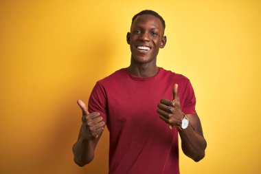 Kırmızı tişörtlü genç Afro-Amerikalı adam izole edilmiş sarı arka plan işareti üzerinde duruyor. Elleriyle olumlu jest yapıyor, gülümsüyor ve mutlu. Neşeli ifade ve kazanan jest.