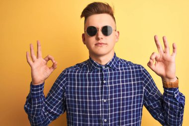 Sarı arka planda günlük gömlek ve güneş gözlüğü takan genç yakışıklı adam rahatla ve parmaklarıyla meditasyon hareketi yaparken gözleri kapalı gülümse. Yoga konsepti.