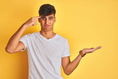 İzole edilmiş sarı arka plan üzerinde duran beyaz tişörtlü genç Hintli adam avuç içinin açık olmasından rahatsız ve şaşırmış bir şekilde fotokopi alanını gösteriyor ve alnını işaret ediyor. Bunu bir düşün..