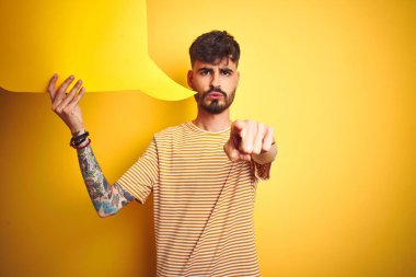 Elinde konuşma balonu tutan dövmeli genç adam izole edilmiş sarı arka planda kamerayı işaret ediyor ve sen, el işareti, ön taraftan pozitif ve kendinden emin bir hareket.