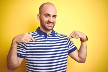 Sakallı genç ve kel bir adam sarı izole edilmiş arka plan üzerine günlük mavi tişört giyiyor. Yüzünde bir gülümsemeyle, gururlu ve mutlu bir şekilde kendini gösteriyor..