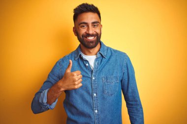 Kotlu tişört giyen genç Hintli adam izole edilmiş sarı arka planda durup mutlu bir el hareketi yapıyor. Kameraya bakıp başarıyı gösteren ifade onaylanıyor.