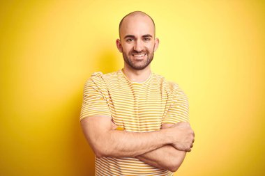 Sarı izole edilmiş arka plan üzerinde sıradan çizgili tişört giyen sakallı genç kel adam kameraya bakarken kollarını kavuşturup gülümsüyor. Pozitif kişi.