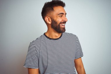Siyah çizgili tişört giyen genç Hintli adam izole edilmiş beyaz arka planda duruyor yüzünde gülümseme, doğal bir ifade ile yan yana bakıyor. Kendinden emin gülen.