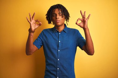 Rastgele kot gömlek giyen Afro bir adam izole edilmiş sarı arka planda dikiliyor gözleri kapalı gülümsüyor parmaklarıyla meditasyon hareketi yapıyor. Yoga konsepti.