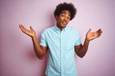 Afro saçlı, mavi tişörtlü, soyutlanmış pembe arka planda duran, kafası karışmış, kolları ve elleri havada genç bir Amerikalı. Şüphe kavramı.
