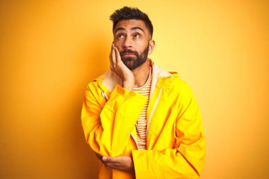 Sarı arka planda duran yağmurluk giyen genç Hintli adam kollarını kavuşturarak depresyondan sıkılmış ve yorgun görünüyor..