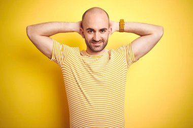 Sakallı genç ve kel bir adam sarı izole edilmiş arka plan üzerine günlük çizgili tişört giyiyor. Rahatlıyor ve esniyor, kolları ve elleri başının ve boynunun arkasında mutlu bir şekilde gülümsüyor.