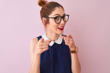 Kırmızı saçlı, atkuyruklu, zarif elbiseli ve gözlüklü pembe arka planda mutlu ve komik yüzlü bir kadın. İyi enerji ve titreşimler.