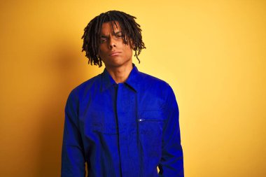 Rastalı, mekanik üniformalı bir Afro işçi izole edilmiş sarı arka plan şüphecisi ve gergin, sorun yüzünden suratını asıyor. Negatif kişi.