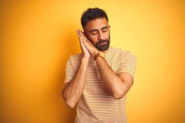 İzole edilmiş sarı arka plan üzerinde duran genç Hintli adam yorgun uyur ve elleri ile birlikte poz verir ve gözleri kapalı gülümserdi..