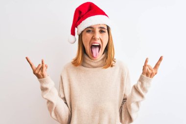 Güzel kızıl saçlı kadın izole edilmiş arka planda noel şapkası takıyor çılgın bir ifadeyle el kaldırarak rock sembolü yapıyor. Müzik yıldızı. Ağır müzik konsepti.