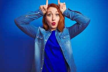 Young beautiful redhead woman wearing denim shirt standing over blue isolated background doing funny gesture with finger over head as bull horns