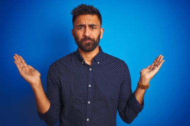 İzole edilmiş mavi arka plan üzerinde duran, kolları ve elleri havada, kafası karışmış genç Hintli zarif bir adam. Şüphe kavramı.