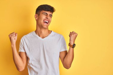 İzole edilmiş sarı arka plan üzerinde duran beyaz tişörtlü genç Hintli adam kolları havada ve gözleri kapalı bir şekilde başarıyı kutluyor ve şaşırıyor. Kazanan konsept.