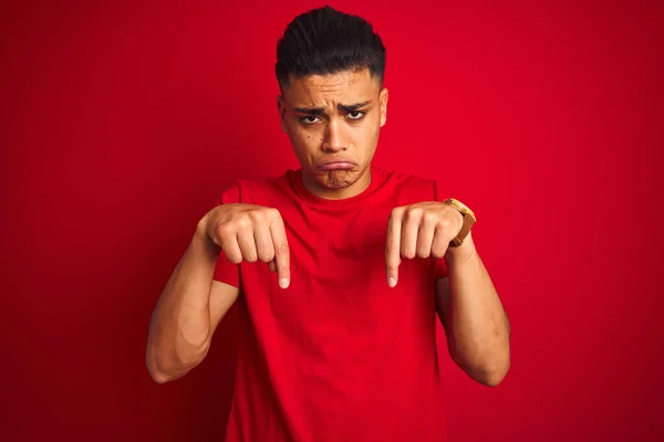 年轻的巴西男子穿着T恤 站在孤立的红色背景上 垂头丧气地垂头丧气 用手指指指着方向 既不快乐又沮丧 — 图库照片