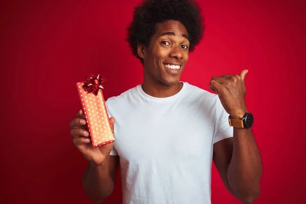 年轻的非洲裔美国人 手里拿着情人节礼物 站在孤立的红色背景上 用大拇指指着侧面 笑容满面 — 图库照片