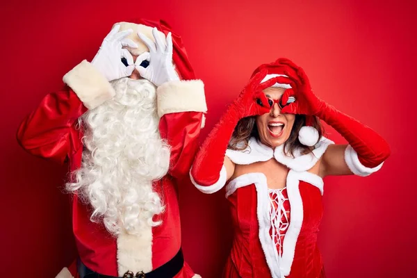 中年夫妇穿着圣诞老人服装 戴着太阳镜 站在孤立的红色背景上 摆出一副像双筒望远镜一样的姿势 伸出舌头 眼睛透过手指看着 疯狂的表情 — 图库照片