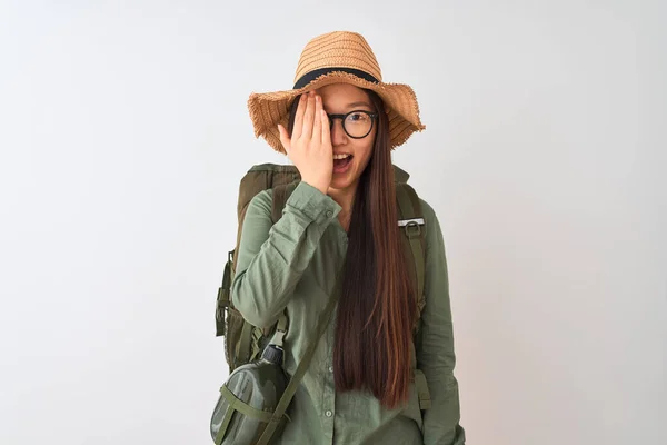 Chinese hiker woman wearing canteen hat glasses backpack over isolated white background covering one eye with hand, confident smile on face and surprise emotion.