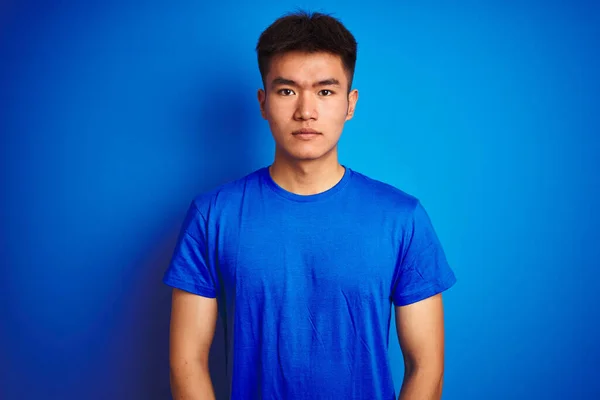 隔離された青い背景の上にTシャツを着た若いアジア系中国人男性が顔に深刻な表情でリラックスしています シンプルで自然なカメラを見て — ストック写真