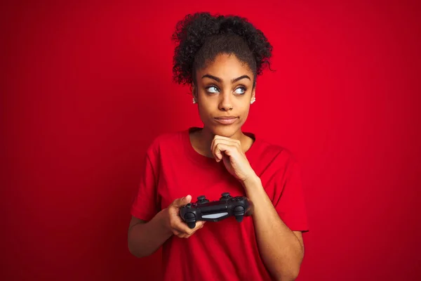 African american gamer woman playing video game using joystick over isolated red background serious face thinking about question, very confused idea