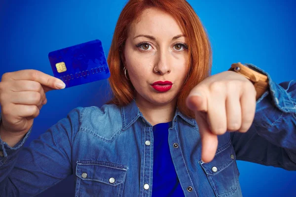 Young beautiful redhead woman holding credit card over blue isolated background pointing with finger to the camera and to you, hand sign, positive and confident gesture from the front