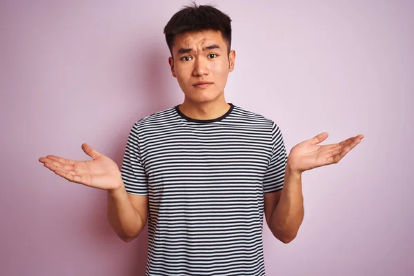 縞模様のTシャツを着た若いアジア系中国人男性が孤立したピンクの背景の上に立っていて 腕や手で上げられた混乱した表情をしています 疑いの概念 — ストック写真