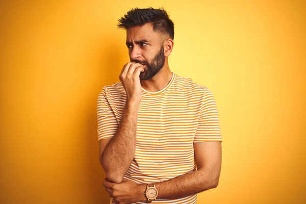 年轻的印度男子穿着T恤站在孤立的黄色背景上 看起来很紧张 双手放在咬指甲的嘴上 焦虑问题 — 图库照片