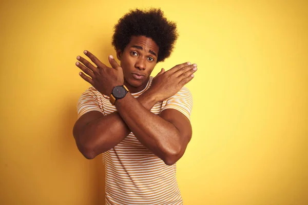 American man with afro hair wearing striped t-shirt standing over isolated yellow background Rejection expression crossing arms doing negative sign, angry face