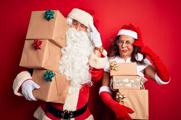 Middle age couple wearing Santa costume holding tower of gifts over isolated red background Shooting and killing oneself pointing hand and fingers to head like gun, suicide gesture.
