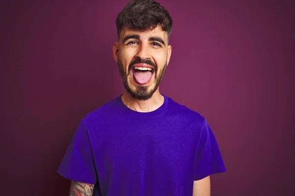有纹身的年轻人穿着T恤 站在孤立的紫色背景上 带着滑稽的表情伸出舌头 情感概念 — 图库照片
